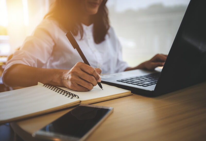 Woman sitting at a desk with left hand on a laptop and right hand with a pen in hand above a notepad.